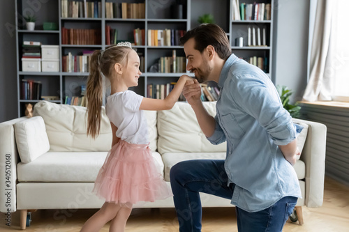 Cute daughter play with father indoor enjoy free time together, dad kneeling holds hand kisses arm of pretty kid girl showing good manners, politeness, gentlemen and little princess having fun at home