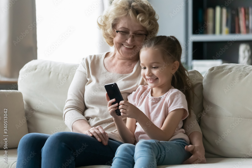In living room sit on sofa resting little granddaughter with old grandma, kid holds cell phone. New generation addicted to wireless gadgets, website content parental control, bad habit overuse concept In living room sit on sofa resting little granddaughter with old grandma, kid holds cell phone. New generation addicted to wireless gadgets, website content parental control, bad habit overuse concept