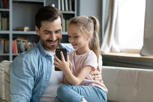 фотографія Small daughter seated on father lap showing him cool game on smartphone in internet