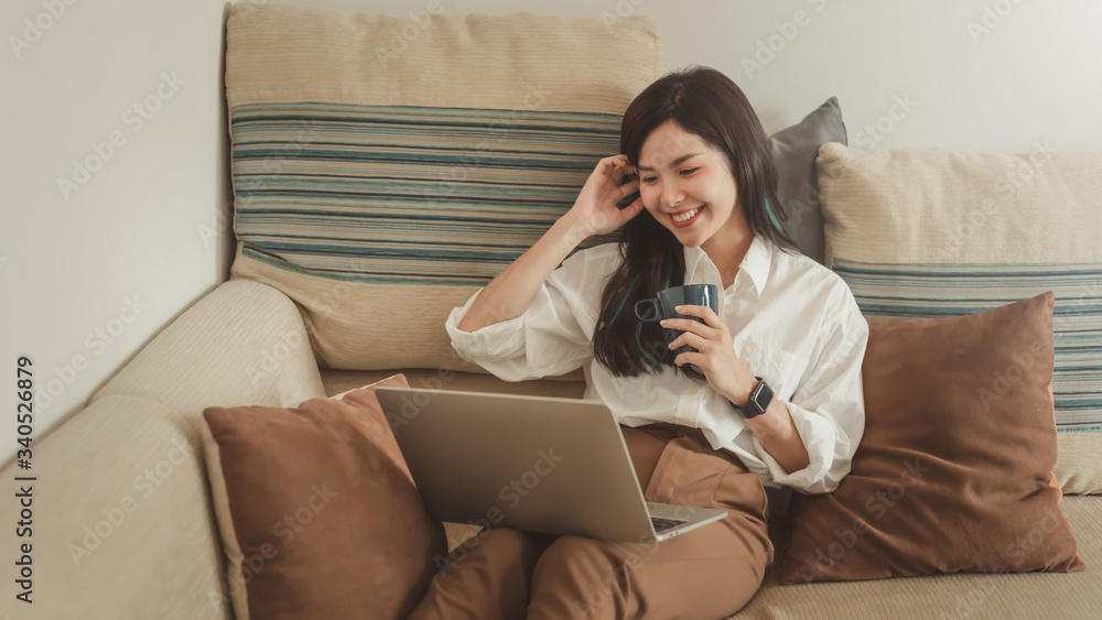 Happy young asian woman with a laptop and drinking coffee sitting on the sofa at home. Happy young asian woman with a laptop and drinking coffee sitting on the sofa at home.