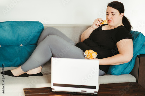 Overeating and gluttony concept. Mindless snacking, home sedentary lifestyle. Obese woman sitting on sofa eating unhealthy food photo