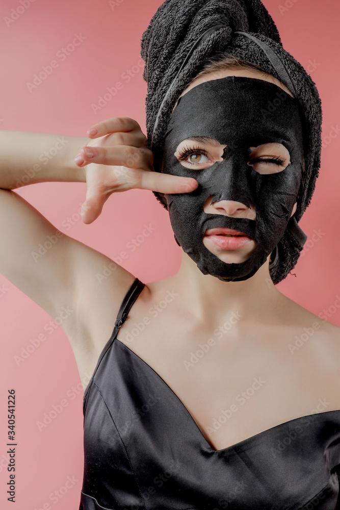 Young woman apply black cosmetic fabric facial mask on pink background. Face peeling mask with charcoal, spa beauty treatment, skincare, cosmetology. Close up Young woman apply black cosmetic fabric facial mask on pink background. Face peeling mask with charcoal, spa beauty treatment, skincare, cosmetology. Close up