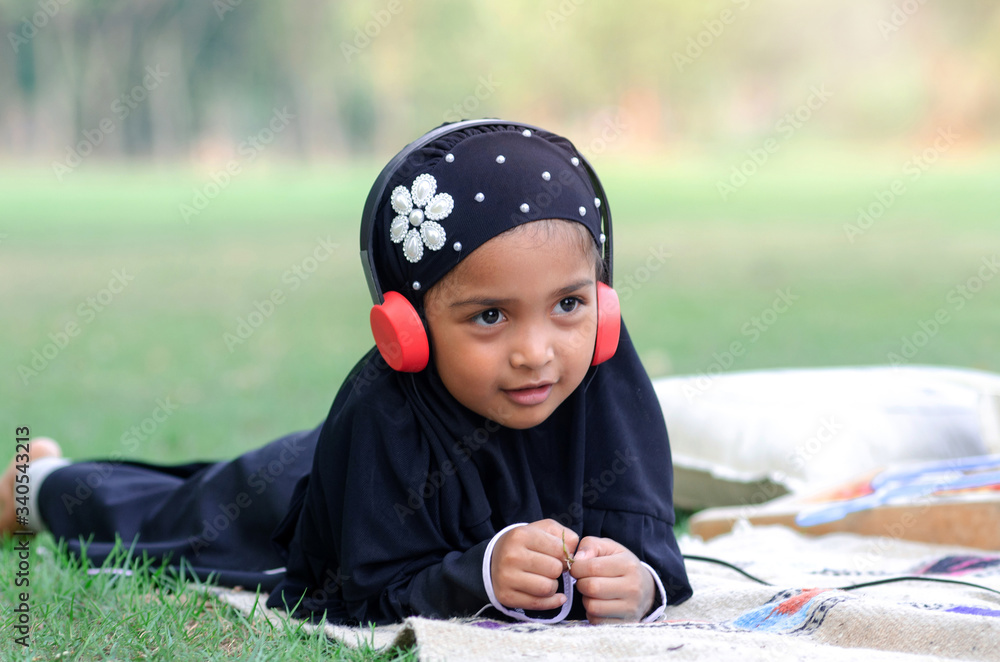 Adorable muslim girl lying on the grass in park, using earphone and wearing black color hijab