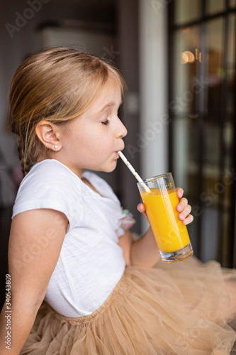 Cute little caucasian girl with blonde hair sitting on the kitchen table and drinking fresh mango juice. Healthy lifestyle concept. Kid 6 years old at home