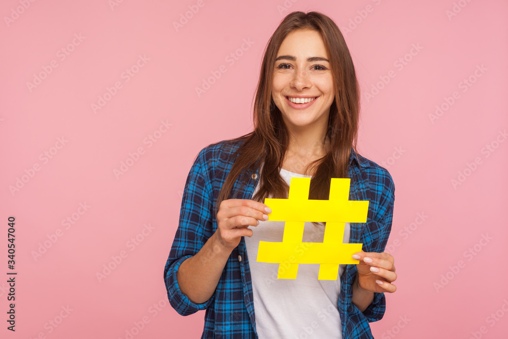 Viral blog post. Happy pretty smiling girl in checkered shirt holding yellow hashtag symbol, making important topic popular, setting trends on Internet. indoor studio shot isolated on pink background Viral blog post. Happy pretty smiling girl in checkered shirt holding yellow hashtag symbol, making important topic popular, setting trends on Internet. indoor studio shot isolated on pink background
