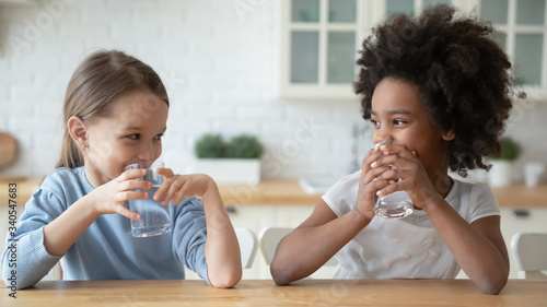 Happy little multiracial sisters sit at table in home kitchen drink pure mineral water, smiling small multiethnic girls children enjoy clean still aqua, feel thirsty, healthy lifestyle concept