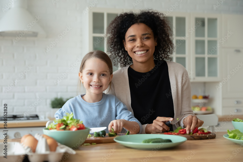 Portrait of smiling African American mom and little Caucasian daughter look at camera cooking in kitchen together, happy biracial mother and small girl child have fun preparing healthy food at home Portrait of smiling African American mom and little Caucasian daughter look at camera cooking in kitchen together, happy biracial mother and small girl child have fun preparing healthy food at home