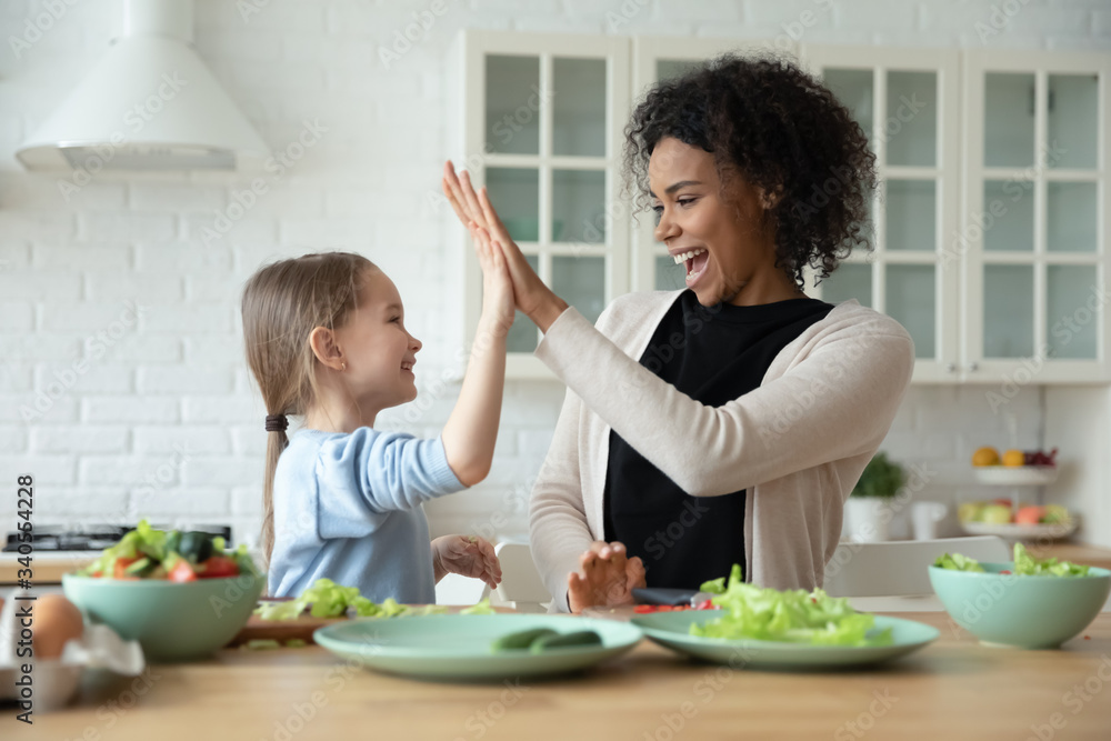 Overjoyed young African American mom and little Caucasian daughter give high five cooking at home together, smiling biracial mother and small girl child have fun preparing healthy food in kitchen Overjoyed young African American mom and little Caucasian daughter give high five cooking at home together, smiling biracial mother and small girl child have fun preparing healthy food in kitchen