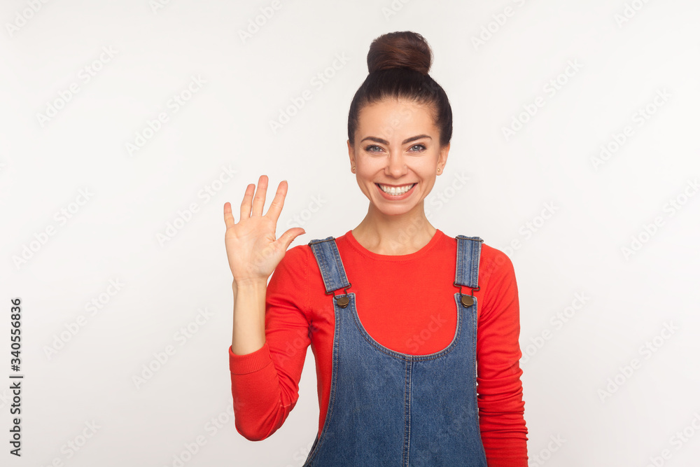 Portrait of friendly stylish pretty girl with hair bun in denim overalls gesturing sweet hello, welcoming with toothy smile, waving hand and greeting. indoor studio shot isolated on white background Portrait of friendly stylish pretty girl with hair bun in denim overalls gesturing sweet hello, welcoming with toothy smile, waving hand and greeting. indoor studio shot isolated on white background