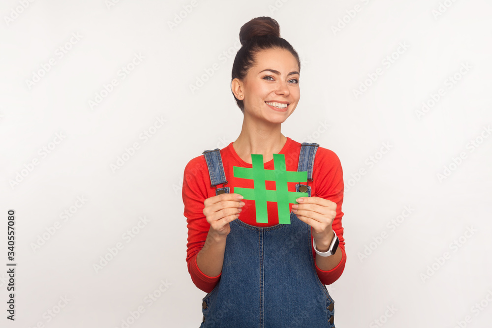 Follow trends, subscribe to popular blog! Portrait of cheerful stylish pretty girl with hair bun in denim overalls holding social media hashtag symbol and smiling. indoor studio shot, white background Follow trends, subscribe to popular blog! Portrait of cheerful stylish pretty girl with hair bun in denim overalls holding social media hashtag symbol and smiling. indoor studio shot, white background