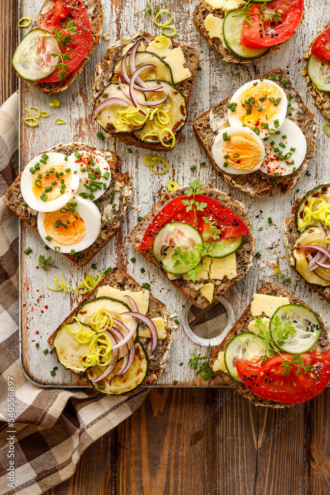 A set of breakfast sandwiches made from homemade buckwheat bread with various toppings on a wooden rustic board, top view. A set of breakfast sandwiches made from homemade buckwheat bread with various toppings on a wooden rustic board, top view.