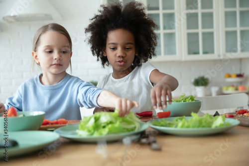 Cute little multiracial girls sisters have fun cooking in modern kitchen together, small preschooler multiethnic children siblings prepare food, make healthy salad for breakfast at home