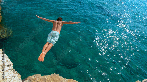 Obraz na plátně COPY SPACE: Man jumping off a rocky ledge and into the glistening blue ocean