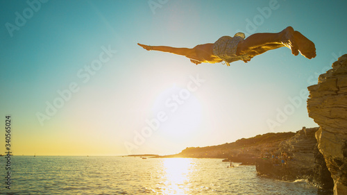 CLOSE UP: Athletic man dives off a cliff and head-first into the ocean at sunset