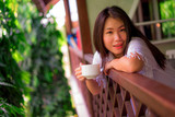 young happy and beautiful Asian Chinese woman having morning coffee or tea at hotel terrace or home balcony leaning on the balustrade relaxed and cheerful
