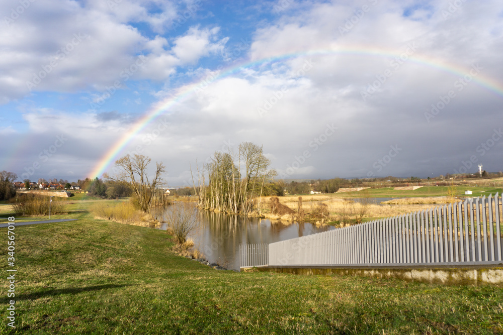 Fluss fließt durch Bäume ohne Blätter hindurch. Regenbogen vor blauem Himmel leuchtet über dem Panorama. Geländer des Staudamms im Bild. Mainaue. Bayreuth. Deutschland.