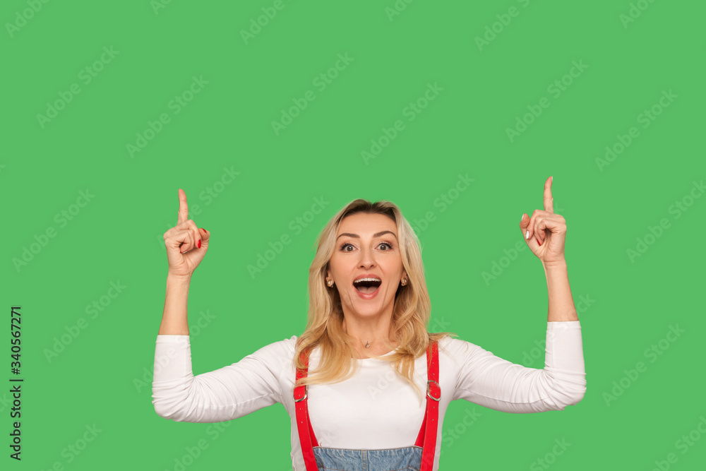 Wow, look at advertise above! Portrait of surprised amazed adult woman in stylish overalls pointing up at blank wall, empty place for promotional message. studio shot isolated on green background Wow, look at advertise above! Portrait of surprised amazed adult woman in stylish overalls pointing up at blank wall, empty place for promotional message. studio shot isolated on green background