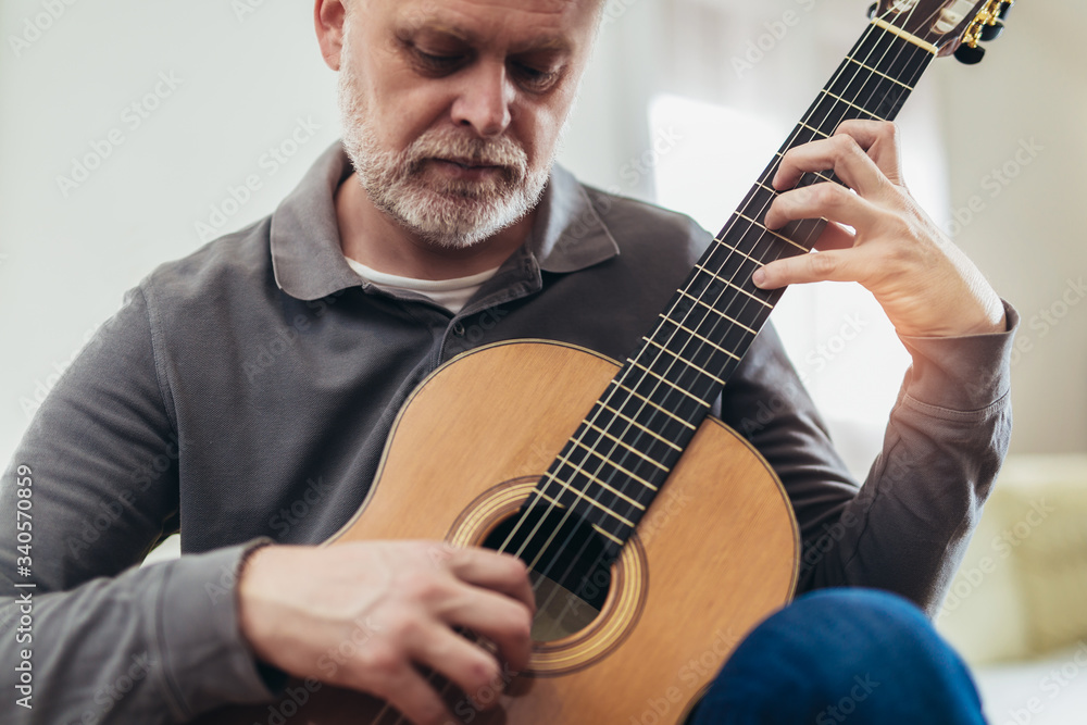 Handsome mature man in casual clothes is smiling while playing guitar at home