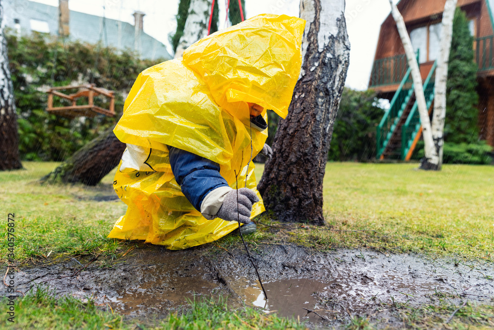 Cute adorable caucasian toddler boy in bright yellow raincoat and wellies playing alone at dirt muddy puddle during cold spring rainy day at home backyard. Carefree childhood concept. Bad weather