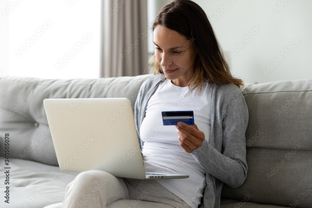 Positive woman paying online, using laptop, looking at screen, shopping in internet store, satisfied young female holding plastic credit card, making purchase, sitting on couch at home Positive woman paying online, using laptop, looking at screen, shopping in internet store, satisfied young female holding plastic credit card, making purchase, sitting on couch at home
