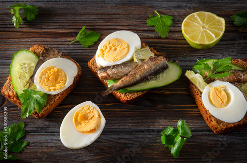 rye bread sandwiches with sprats, fresh cucumber, parsley, lime and boiled egg on a dark wooden background, top view, canned fish,