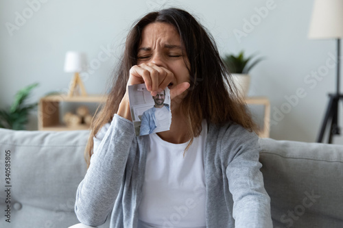 Depressed young woman crying, holding boyfriend or husband picture close up, stressed upset girl suffering from break up or divorce, sitting on couch at home alone, feeling lonely and hopeless