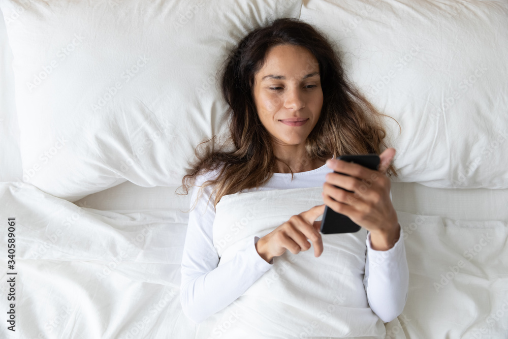 Top view woman using phone, relaxing in comfortable bed in morning close up, checking social network, reading message, beautiful girl looking at smartphone screen, lying on soft pillow in bedroom Top view woman using phone, relaxing in comfortable bed in morning close up, checking social network, reading message, beautiful girl looking at smartphone screen, lying on soft pillow in bedroom