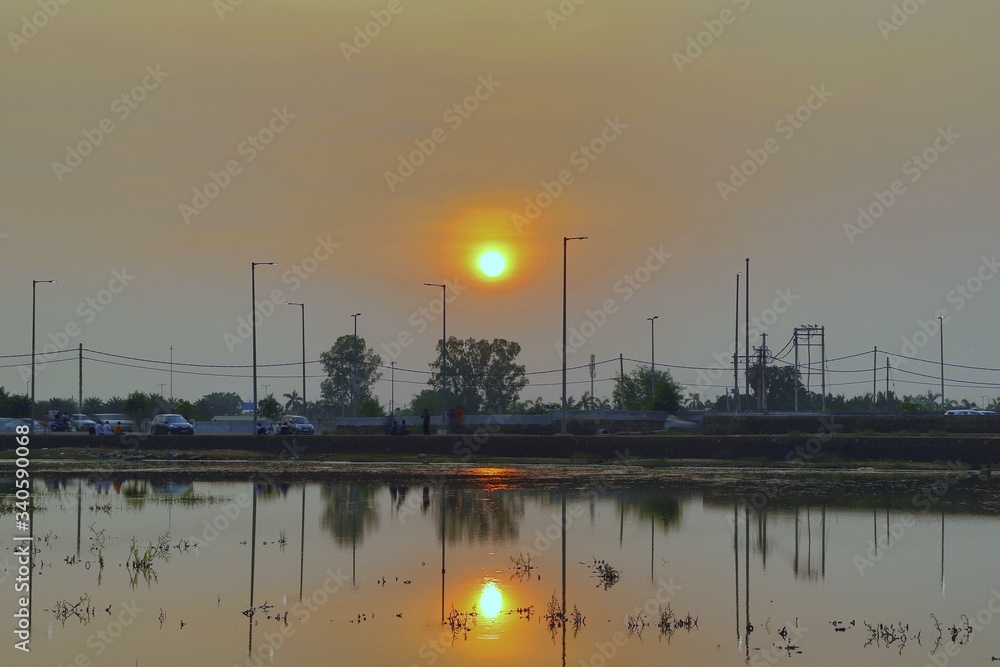 Sunset beyond the pond and Trees, power lines and power poles reflected in a water, making  lines
