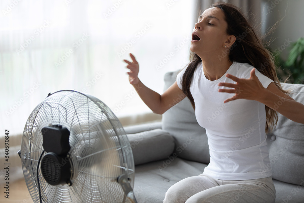 Overheated woman enjoying fresh air, cooling herself by electric fan close  up, exhausted tired pretty girl sitting on couch in front of ventilator,  suffering from hot summer weather at home Photos