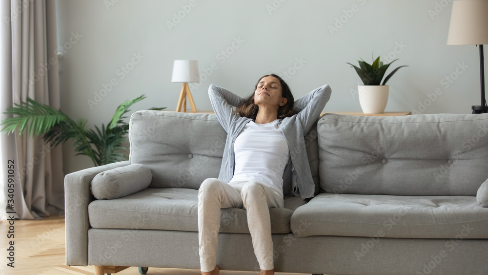 Peaceful young woman with hands behind head relaxing on cozy sofa at home, daydreaming and meditating, satisfied serene girl leaning back, stretching on comfortable couch in living room