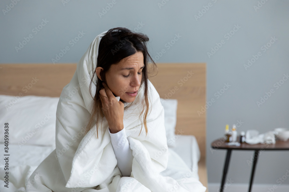 Sick young woman wrapped suffering from cold symptoms, sitting on bed at home, unhealthy girl