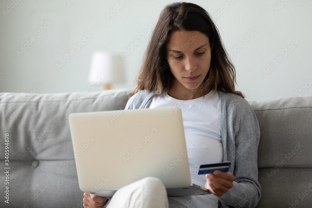 Serious woman holding credit card, reading information, using laptop, sitting on sofa at home, young female customer shopping online in internet store, making purchase, digital payment or banking Serious woman holding credit card, reading information, using laptop, sitting on sofa at home, young female customer shopping online in internet store, making purchase, digital payment or banking