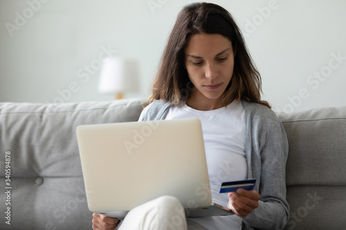 Serious woman holding credit card, reading information, using laptop, sitting on sofa at home, young female customer shopping online in internet store, making purchase, digital payment or banking © fizkes