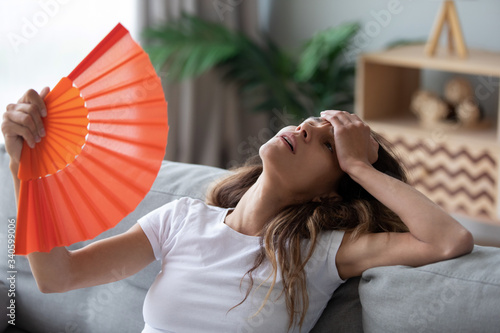 Tela Overheated woman sitting on couch, waving orange paper fan close up, girl feelin