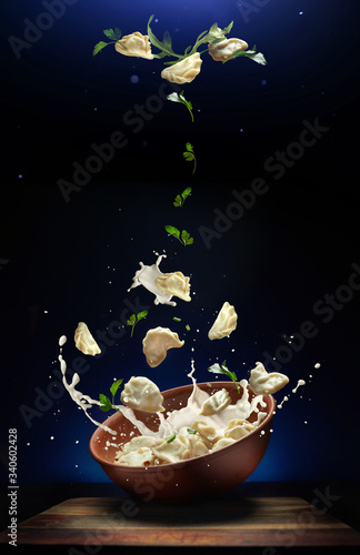 Pierogi flying out of the clay bowl with cream and parsley. Some vareniki stay inside the plate. Blue background. Ornamented with levitating ingredients. 