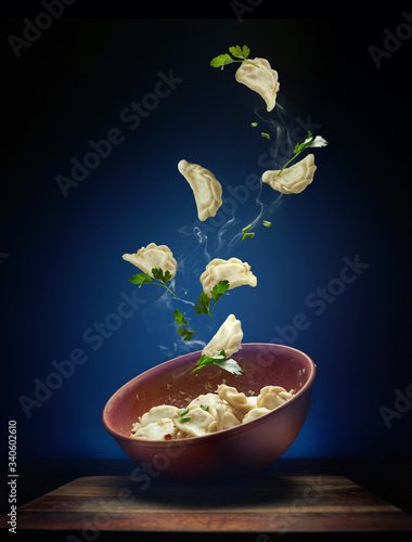 Hot pierogi flying out of the clay bowl with cream and parsley. Some vareniki stay inside the plate.  Blue background.