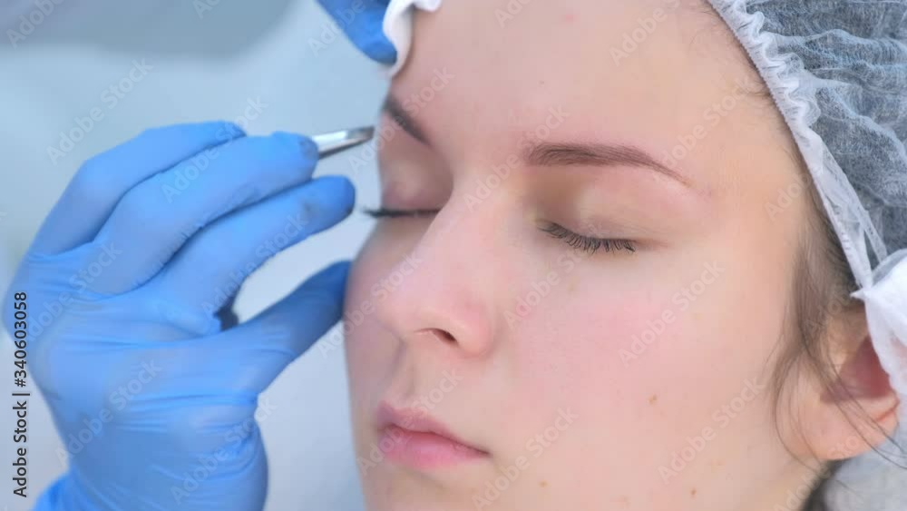 Cosmetologist plucks out hair on eyebrows with tweezers for woman, face closeup. Eyebrows tweezing procedure. Beautician is making brows correction with cosmetic tweezer. Beauty industry. Cosmetologist plucks out hair on eyebrows with tweezers for woman, face closeup. Eyebrows tweezing procedure. Beautician is making brows correction with cosmetic tweezer. Beauty industry.