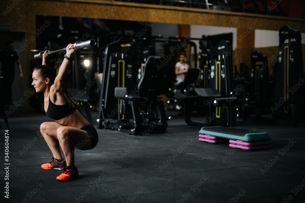 Strong young woman with perfect athletic body wearing black sportswear lifting empty vulture from the barbell overhead during sport workout training in modern dark gym. Concept of healthy lifestyle. Strong young woman with perfect athletic body wearing black sportswear lifting empty vulture from the barbell overhead during sport workout training in modern dark gym. Concept of healthy lifestyle.