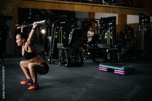 Strong young woman with perfect athletic body wearing black sportswear lifting empty vulture from the barbell overhead during sport workout training in modern dark gym. Concept of healthy lifestyle.