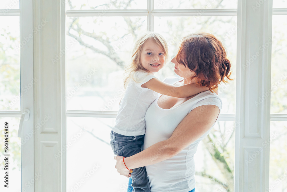 Stay Home Stay Safe. Young mother holding her child. Woman and little girl relaxing in white bedroom near windiow indoors. Happy family at home. Young mom playing with her daughter.