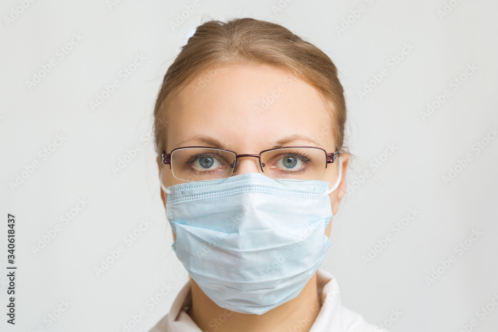 Young female doctor with glasses isolated on light gray background. Respirator mask for mouth and nose covering in virus quarantine time. First protection. Healthcare concept. Closeup. Front view. Young female doctor with glasses isolated on light gray background. Respirator mask for mouth and nose covering in virus quarantine time. First protection. Healthcare concept. Closeup. Front view.