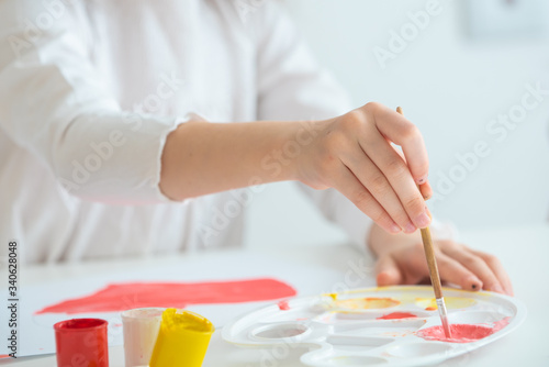 child with a brush mixes pink paint in a palette on a white table with a palette