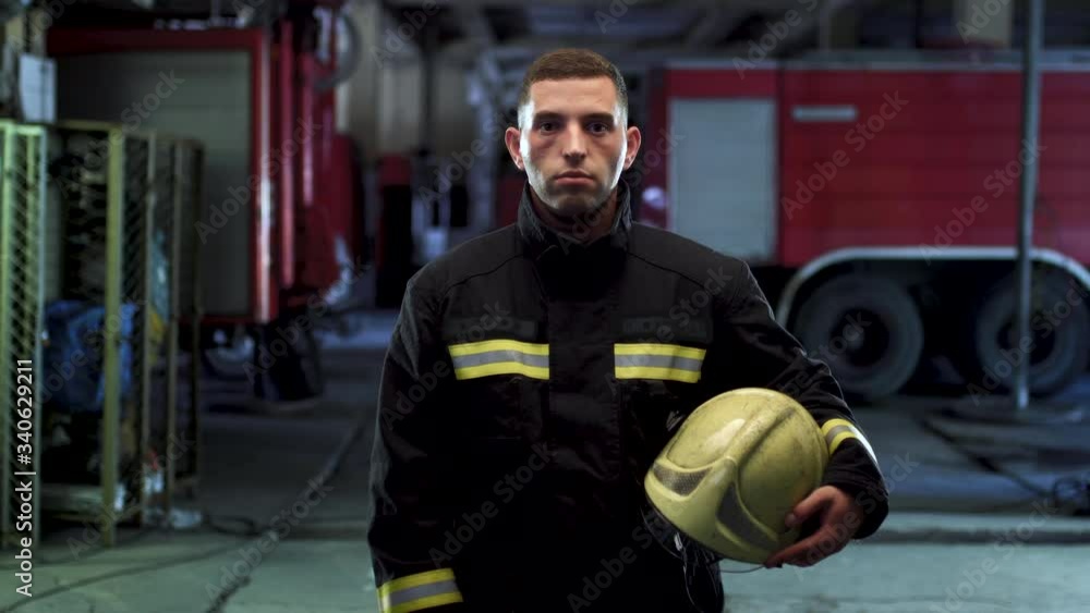 Firefighter video portrait wearing uniform and holding yellow protective helmet. Fire trucks in the background. Camera moving left. Firefighter video portrait wearing uniform and holding yellow protective helmet. Fire trucks in the background. Camera moving left.