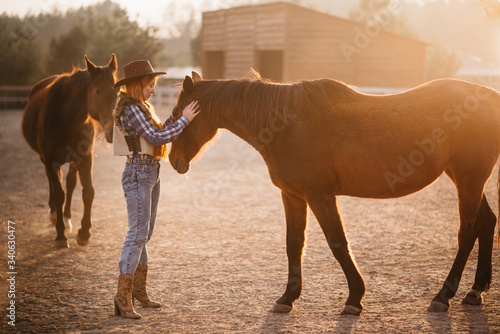 Young woman farmer stroking a brown horse in a corral on a ranch at sunset. Flare effect. © MZaitsev