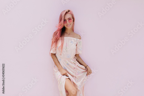 Carefree girl in stylish summer dress posing with interest smile. Inspired caucasian woman with pink hair standing on white background.