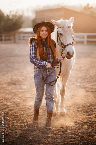 A young woman farmer leads a horse in a corral on a ranch at sunset in the backlight. Full-length portrait.