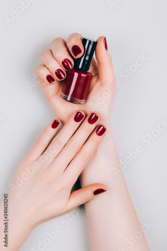 Beautiful dark red manicure with a bottle of nail polish in hands on a grey background. Procedures concept. Flat lay style.