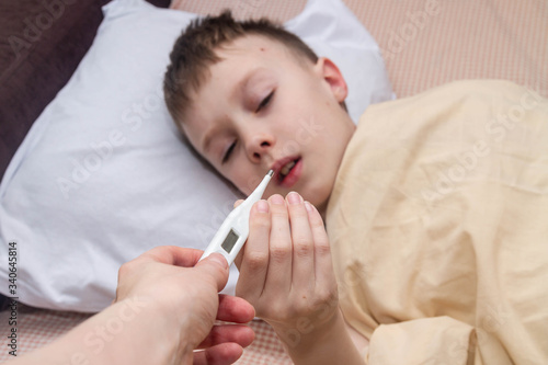 A teenage European boy is lying on the bed. The child is ill. The boy takes a thermometer to measure his body temperature.