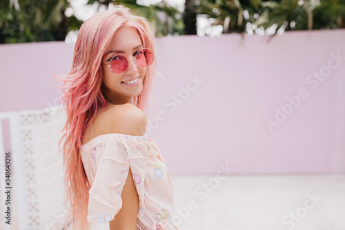 Cute woman with tanned skin looking over shoulder on street background and laughing. Fashionable female model with pink hair enjoying good summer weather. photo