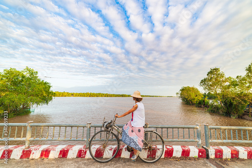 Tourist riding bicycle in the Mekong Delta region, Ben Tre, South Vietnam. Woman having fun cycling among green tropical woodland and water canals. Side view sunset dramatic sky.